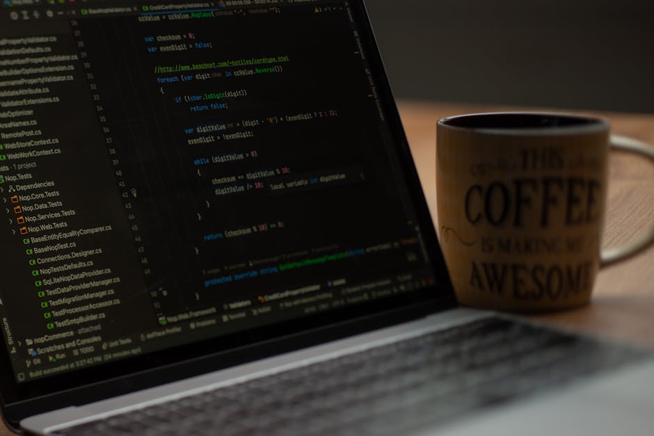 A close-up view of a laptop computer displaying lines of colorful code on a dark background, situated on a wooden desk. Next to the laptop, there is a ceramic coffee mug with a beige background and bl